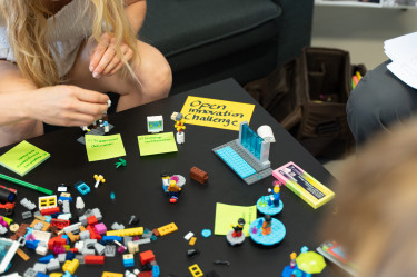 Aus einem Tisch liegen Postits und Legosteine. Man sieht zwei Hände, die gerade etwas bauen und einen Zettel auf dem 