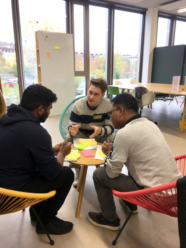 Drei junge Männer sitzen auf bunten Sesseln an einem runden Tisch und diskutieren. Im Hintergrund sind Stellwände und Büromöbel des CoCreationLabs zu sehen.
